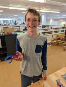 A smiling boy with glasses stands indoors, holding a small robotic claw made of red and blue parts. He is wearing a gray shirt with blue sleeves and a blue pocket. Office supplies and shelves are visible in the background.