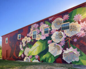 A large mural painted on the side of a brick building features white and pink flowers with green leaves. Three rectangular windows are visible above the mural. The sky is clear and blue.
