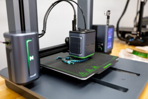 A 3D printer is actively printing an object, with part of a blue and black item visible on the build plate. The machine shows indicator lights and has a touchscreen interface on the right side.