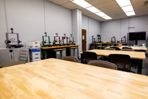 A classroom with several wooden tables, black chairs, and a row of 3D printers in various colors along the wall. There are also boxes, cabinets, and a large monitor in the background.