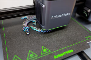 A 3D printer labeled AnkerMake is printing a colorful, low-poly lizard model on a textured print bed with green markings and AnkerMake branding.