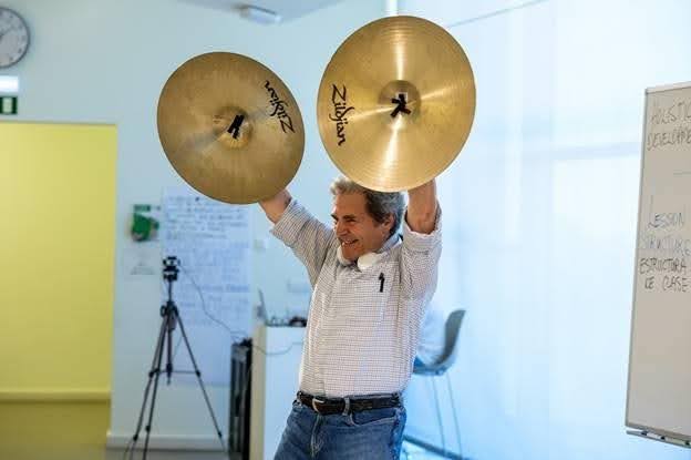 A man in a checked shirt stands indoors holding two large Zildjian cymbals above his head, smiling. There is a whiteboard, tripod, and papers visible in the background.