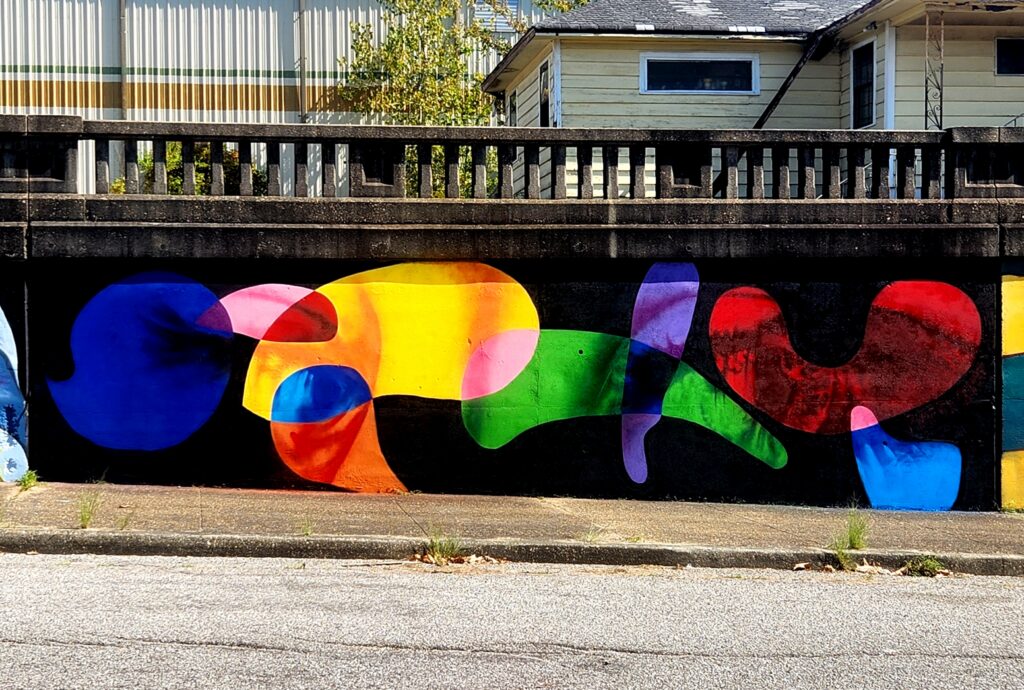 A mural on a concrete wall featuring abstract, colorful shapes in blue, orange, yellow, pink, red, and green. The wall is beside a sidewalk, with a house and trees visible in the background.
