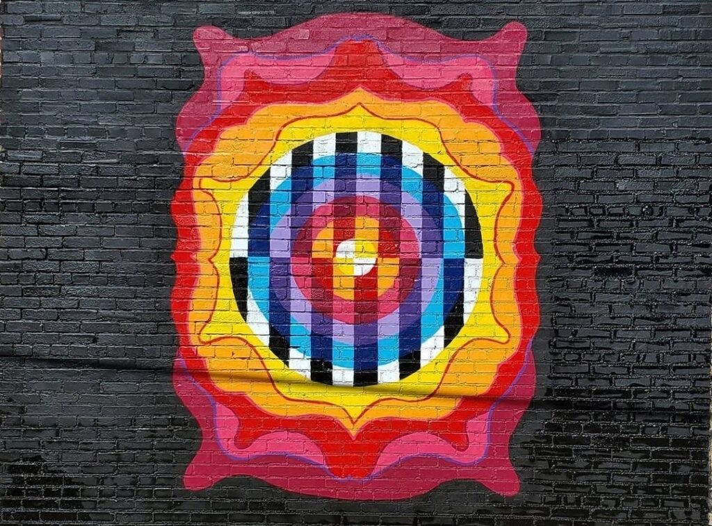 A mural on a brick wall features a colorful geometric design with concentric shapes in red, orange, and yellow. The center contains black and white vertical stripes and blue accents arranged in circular patterns.