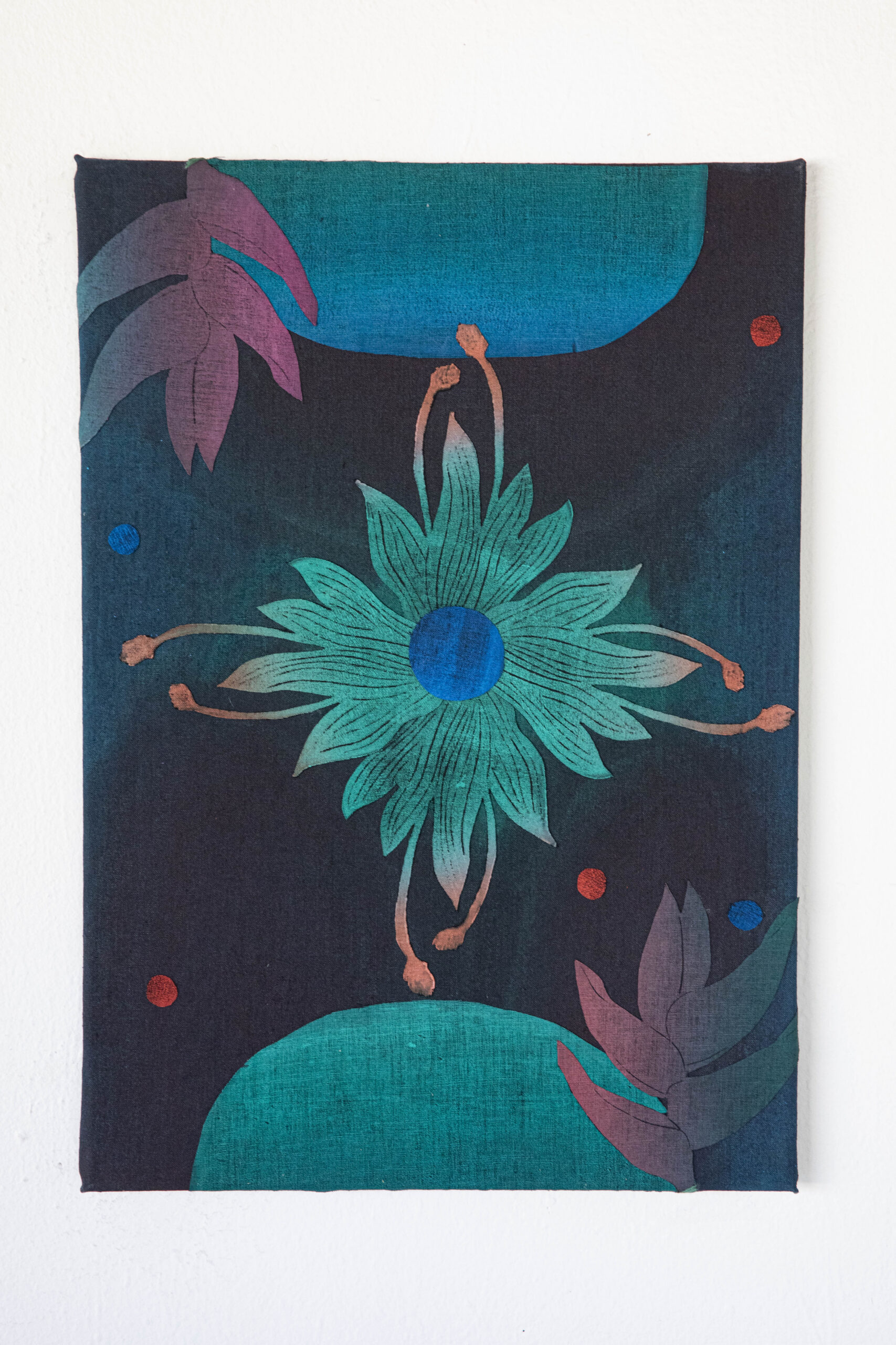 A fabric artwork displays a green and blue stylized flower at the center, with pinkish leaves and red and blue dots on a black background. The design is symmetrical and abstract, with curved teal shapes at the corners.
