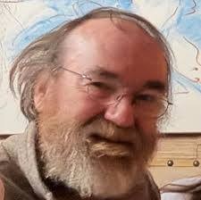 An older man with thinning gray hair, glasses, and a full beard is facing the camera and smiling. The background is blurred, showing a painting with blue and beige tones.