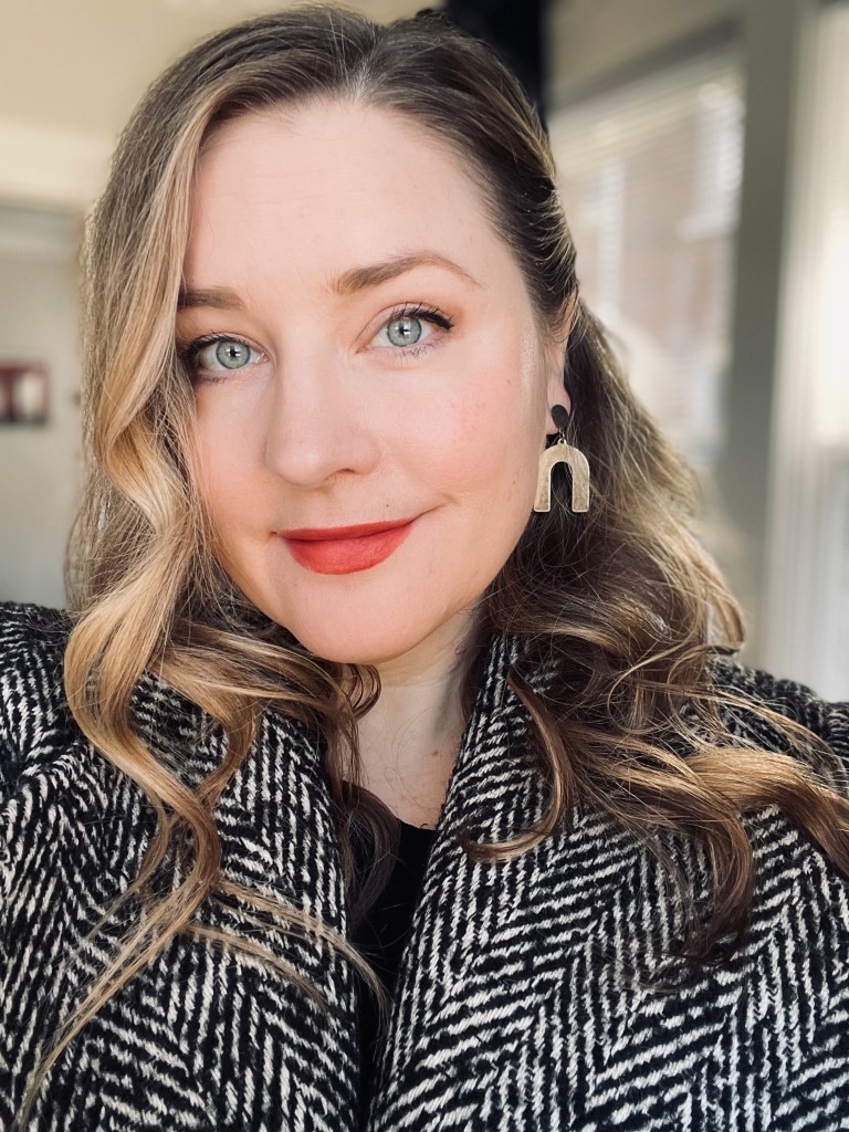 A woman with wavy blonde hair, light eyes, and red lipstick is wearing a patterned coat and statement earrings. She is looking at the camera and smiling slightly. Blurred indoor background with sunlight through windows.