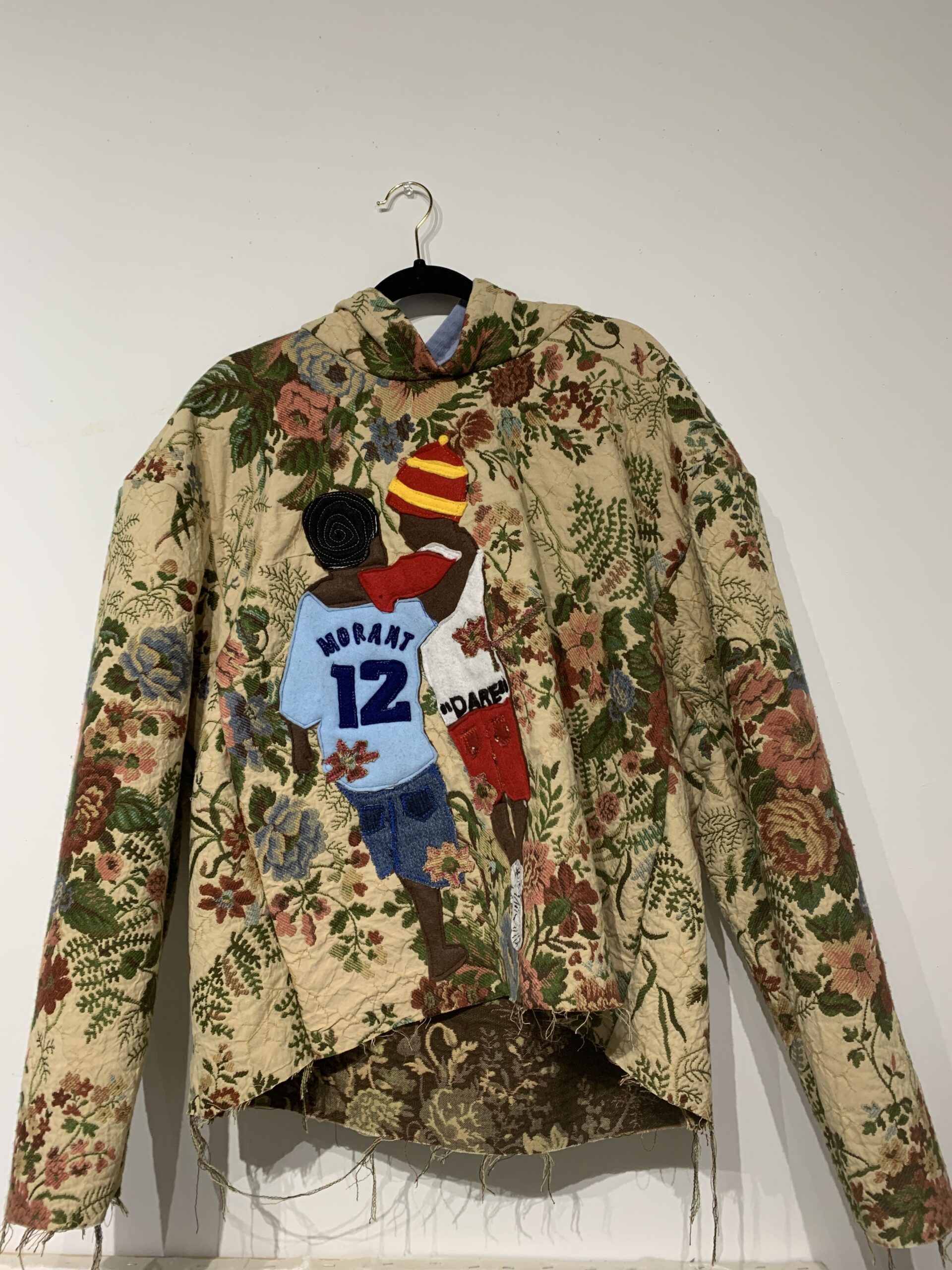 A floral-patterned jacket hangs on a black hanger against a white wall. The back features two appliqué figures, one wearing a blue Morant 12 jersey and the other in red, both appearing to play basketball.