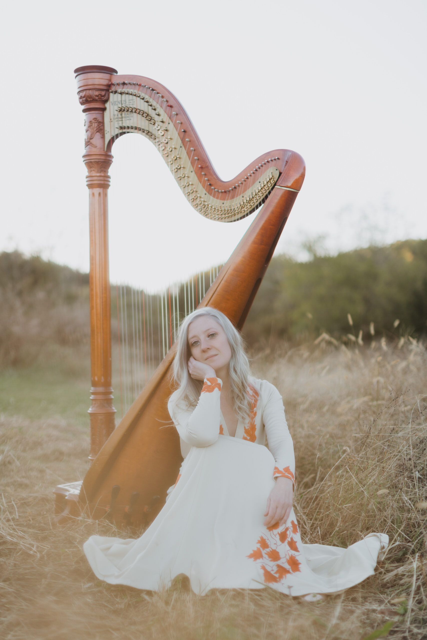 A woman in a white dress with orange flowers sits on the ground in front of a large wooden harp in an outdoor field with tall grass and greenery in the background.