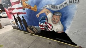 A mural on a sidewalk wall depicts a man flexing his arm, holding a guitar with an American flag design, an airplane, and silhouettes of soldiers. A banner reads Welcome to Aaron Tippin Alley..
