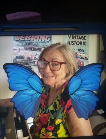 An older woman with glasses and a colorful floral shirt smiles at the camera. Blue butterfly wings are digitally added to her back. Vintage car posters are visible on the wall behind her.
