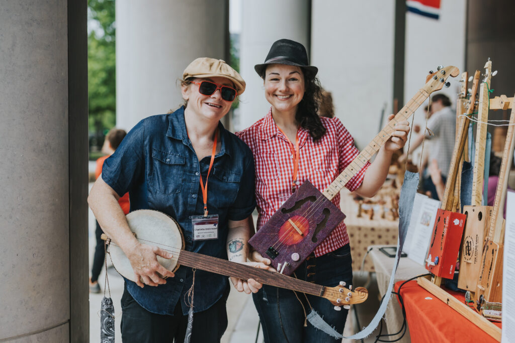 Two people stand side by side, smiling at the camera while holding handmade stringed instruments. They are at an outdoor market or fair with tables and other instruments visible in the background.