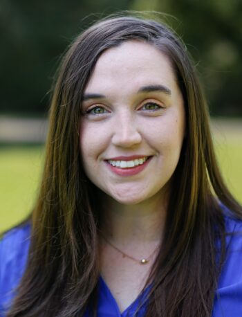 A woman with long brown hair and a blue blouse is standing outdoors, smiling at the camera. The background is blurred greenery and sunlight.