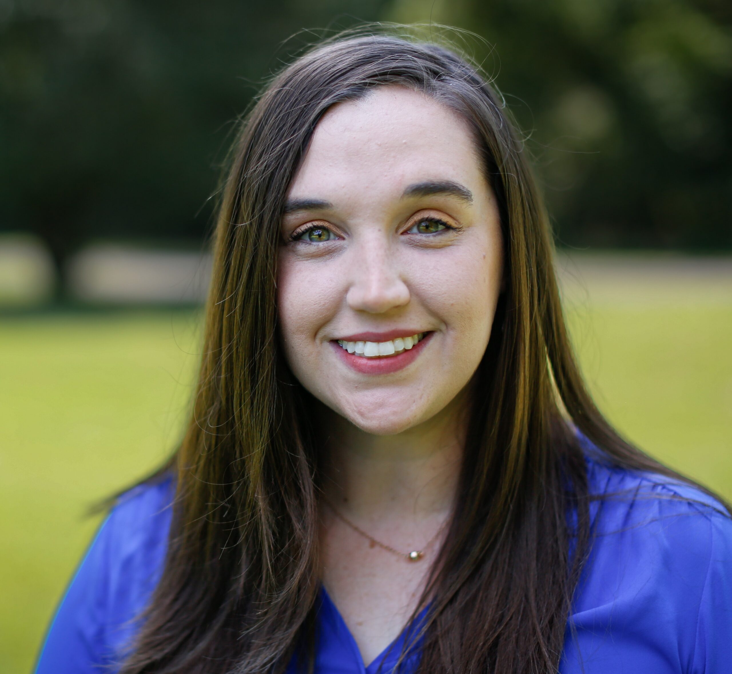 A woman with long brown hair and a blue blouse is standing outdoors, smiling at the camera. The background is blurred greenery and sunlight.