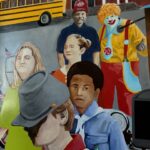 A painting shows a school bus, a building, and six people, including a clown in bright costumes, a man in a red hat, and several children. The scene is colorful and detailed, with various facial expressions and outfits.