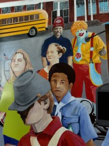 A painting shows a school bus, a building, and six people, including a clown in bright costumes, a man in a red hat, and several children. The scene is colorful and detailed, with various facial expressions and outfits.