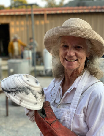 An older woman in a straw hat and white shirt smiles while holding a pottery piece with black markings. She wears a red glove on one hand. A building and another person are visible in the background.