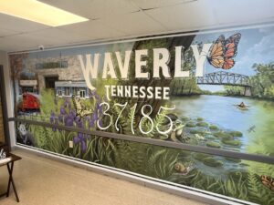 A mural shows Waverly Tennessee 37185 with local scenery: a train, a house, purple flowers, a bridge, water with lily pads, a person in a canoe, butterflies, turtles, and trees under a blue sky.