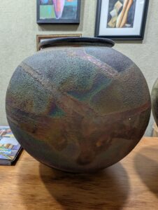 Large, round ceramic pot with a rough, textured surface and subtle earthy colors sits on a wooden table. Framed abstract artwork hangs on the beige wall in the background. A magazine is partially visible on the left.