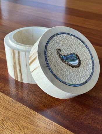 A small, round wooden box with an open lid featuring a textured inlay of a paisley shape made from abalone shell and a border of blue shell pieces.