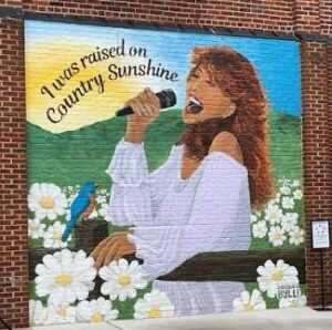 Mural on a brick wall featuring a woman singing into a microphone with the text I was raised on Country Sunshine above her, surrounded by white daisies and a small blue bird.